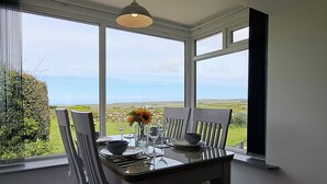 Une table à manger avec des chaises, un vase rempli de fleurs et une vue sur la mer et le paysage, visible à travers de grandes fenêtres.