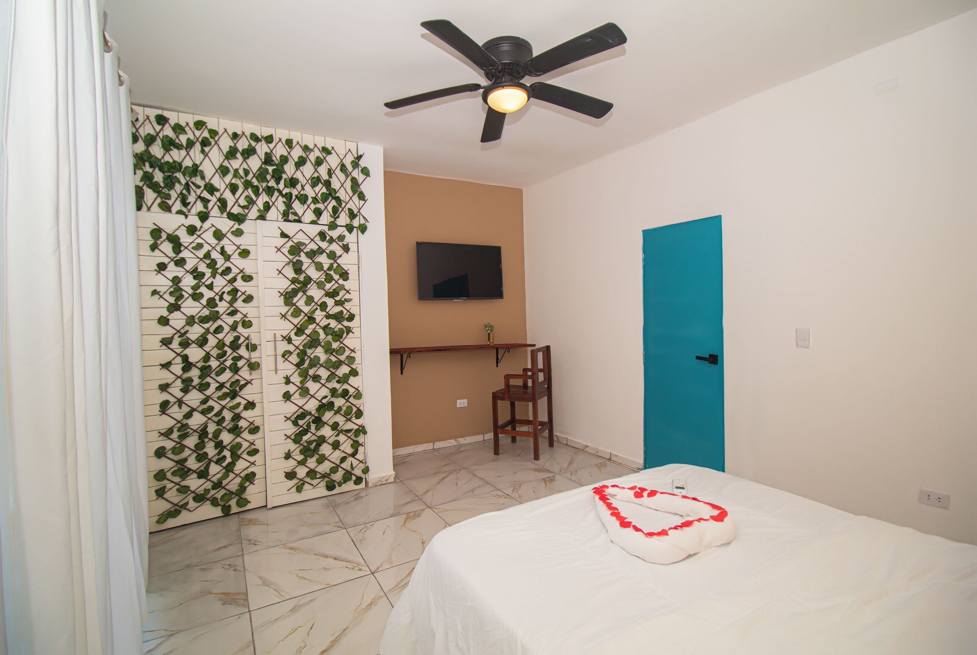 A hotel room with a bed, a ceiling fan, a TV mounted on the wall, a blue door, and a chair.