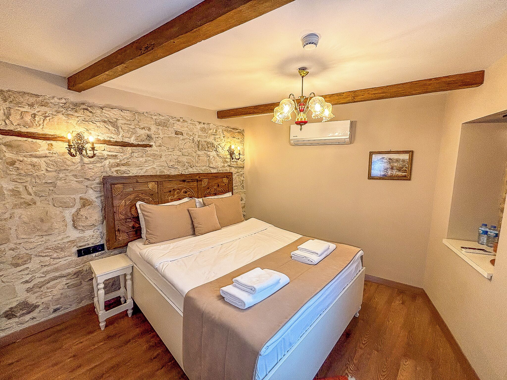 Een slaapkamer met een stenen muur, een houten balk in het plafond, een bed met hoofdeinde, bedlampen en een plafondarmatuur.