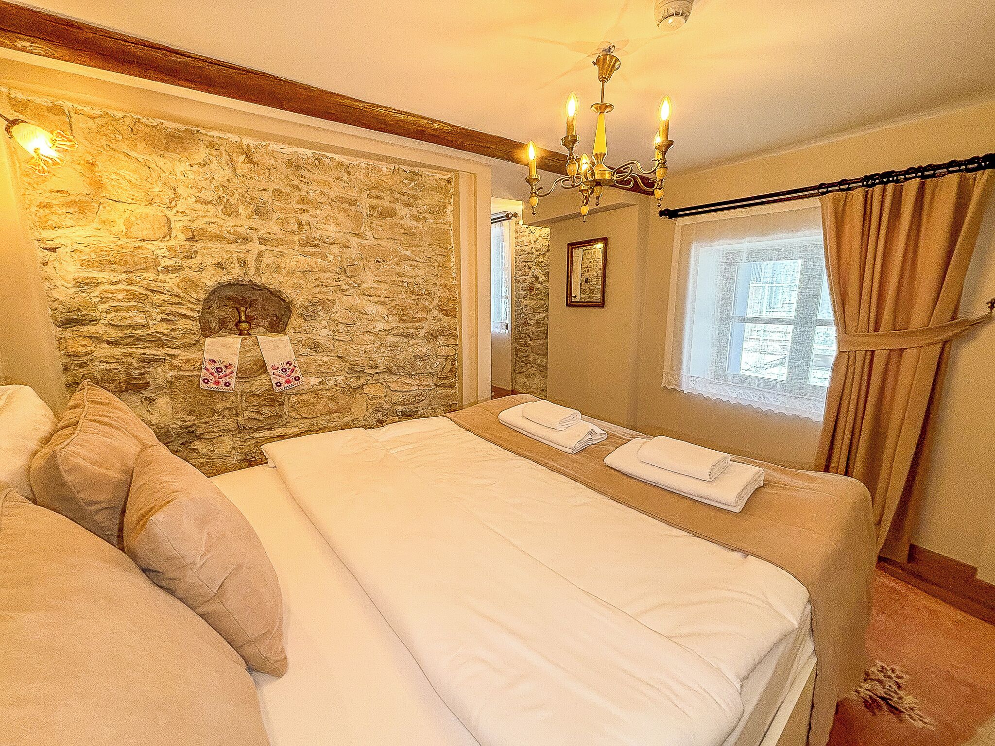 Un dormitorio con pared de piedra, una araña, cama con ropa blanca y una ventana con cortinas.