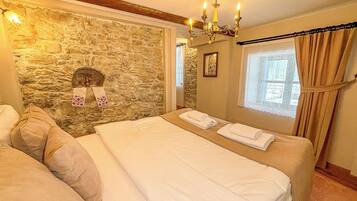 Un dormitorio con pared de piedra, una araña, cama con ropa blanca y una ventana con cortinas.