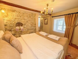 Un dormitorio con pared de piedra, una araña, cama con ropa blanca y una ventana con cortinas.