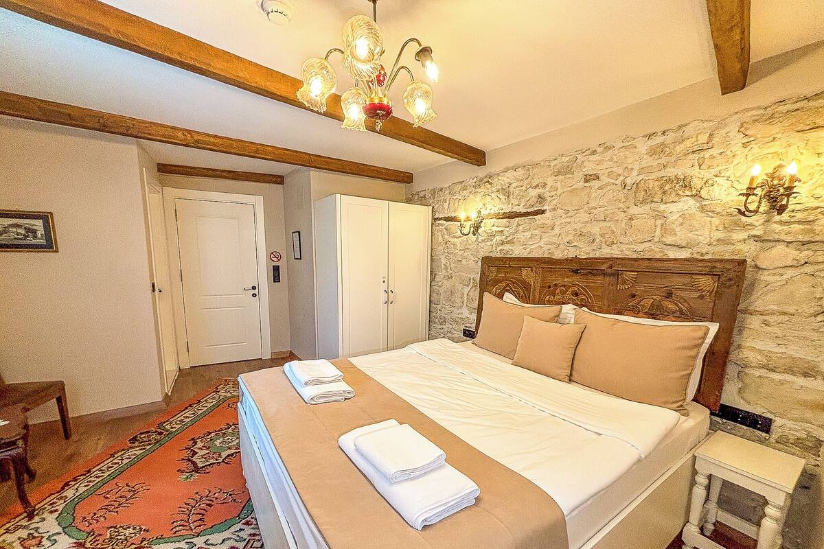 A bedroom with a bed, a nightstand, a chair, a wooden ceiling, and a stone wall.