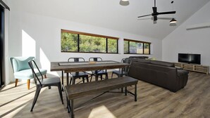 A modern living room with a dining area, large windows, a ceiling fan, and a flat-screen TV.