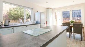 A modern kitchen with a large island, white cabinetry, and a view of the backyard through large windows. - Sunnyside (Wells-next-the-Sea)