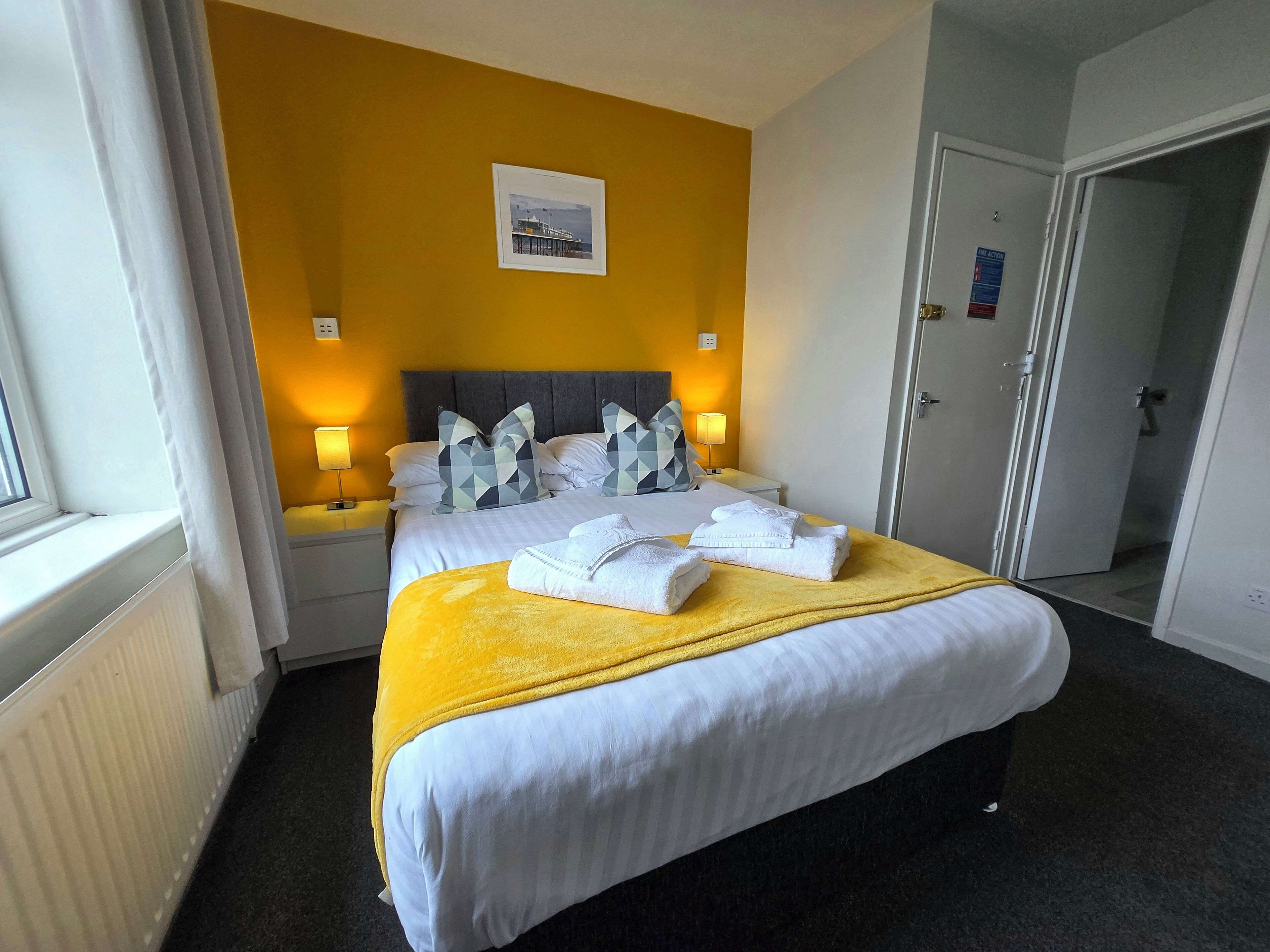 A hotel room with a bed, two bedside tables, a nightstand, a window with curtains, a picture on the wall, and a door.