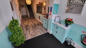 A hallway with a wooden floor, a reception desk with a 'hello' sign, a mirror, and a staircase leading to another room. - Beach Hotel Torbay (Paignton)