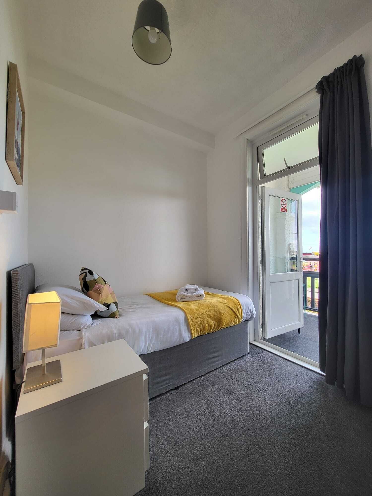 Una habitación de hotel con una cama, una mesita de noche con lámpara, un armario pequeño y un ventanal corredero al exterior.