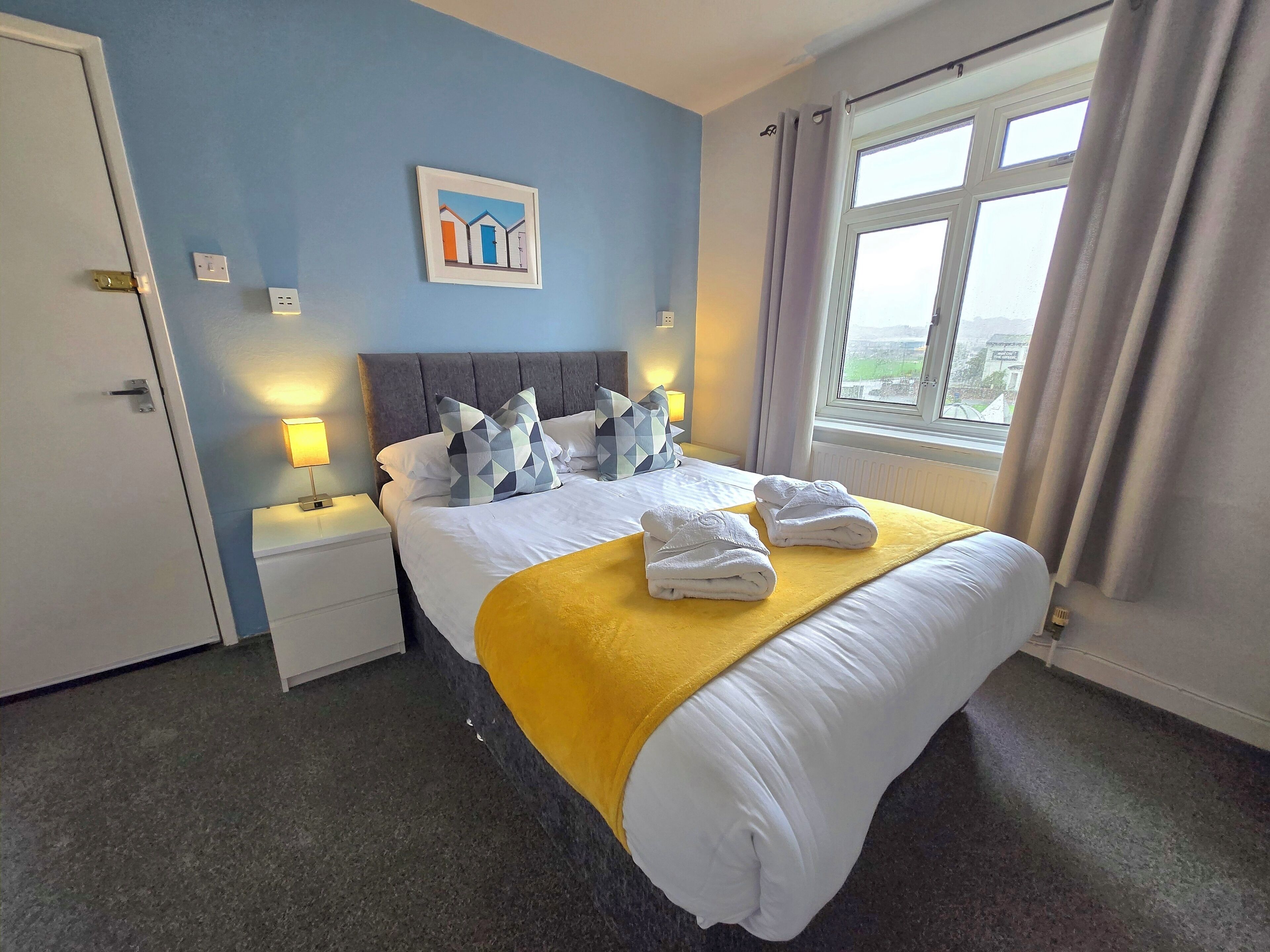 A hotel room with a bed, a nightstand, a lamp, a window with curtains, and a painting on the wall.