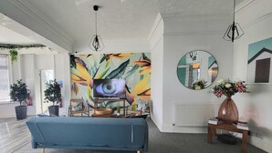 A modern living room with a blue sofa, a television, a large abstract mural, a round mirror, and a vase with flowers. - Beach Hotel Torbay (Paignton)