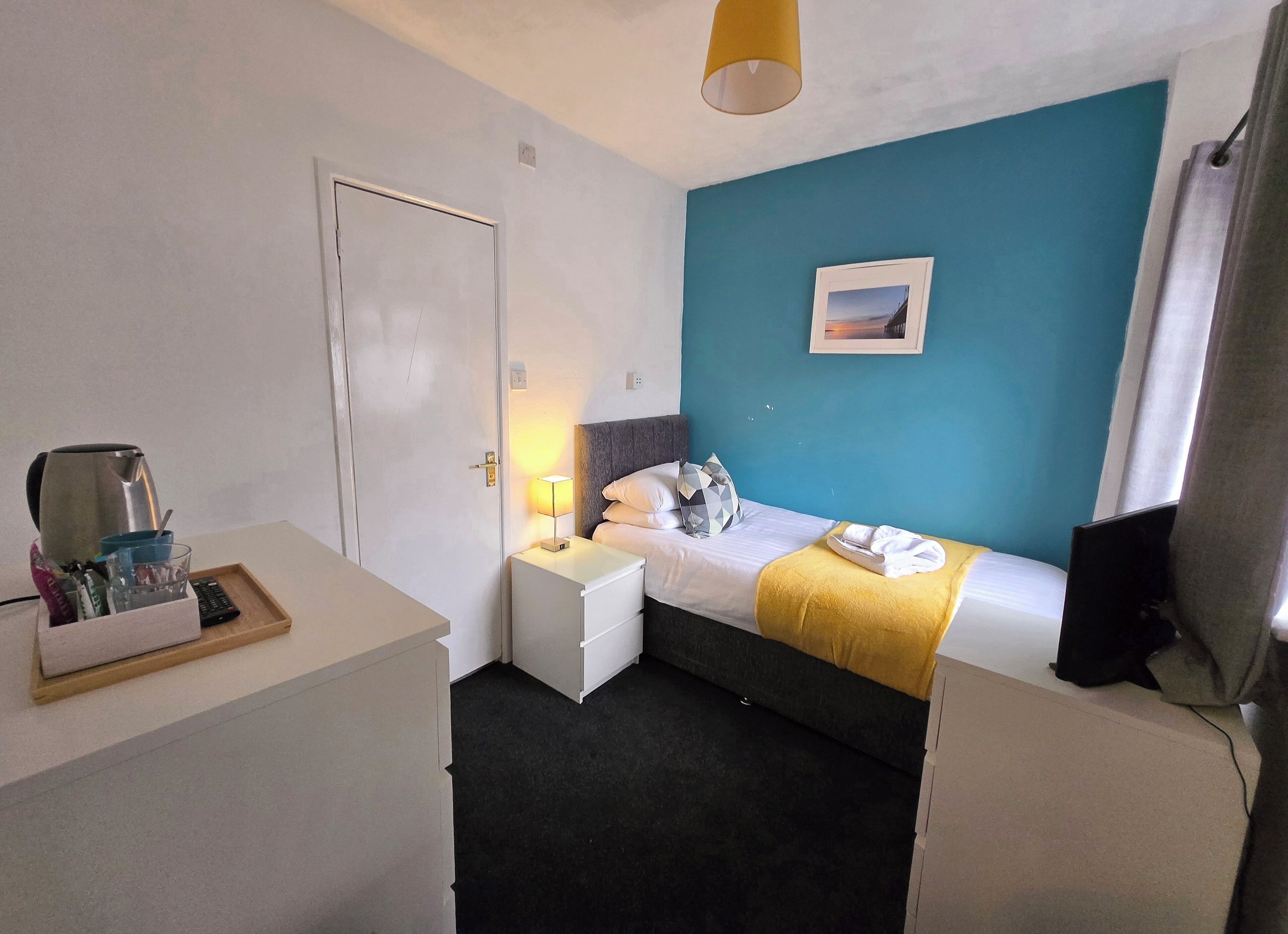 Een hotelkamer met een bed, nachtkastje, lamp, foto aan de muur en televisie op een standaard.