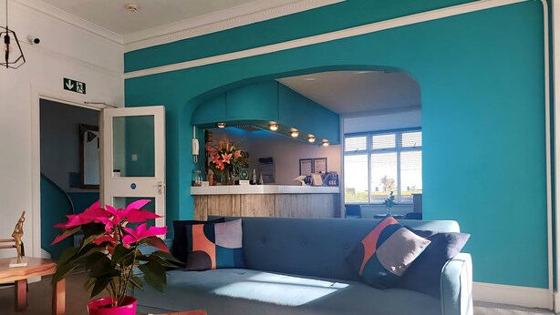 A modern living room with a teal accent wall, a blue sofa, a wooden coffee table with a potted plant, and a reception desk with a mirror and flowers. - Beach Hotel Torbay (Paignton)