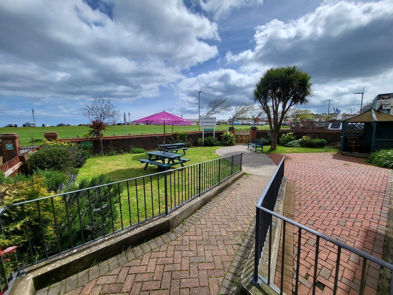 A paved walkway with a metal railing, a picnic table, and a tree in a landscaped area with a grassy field and a building in the background.