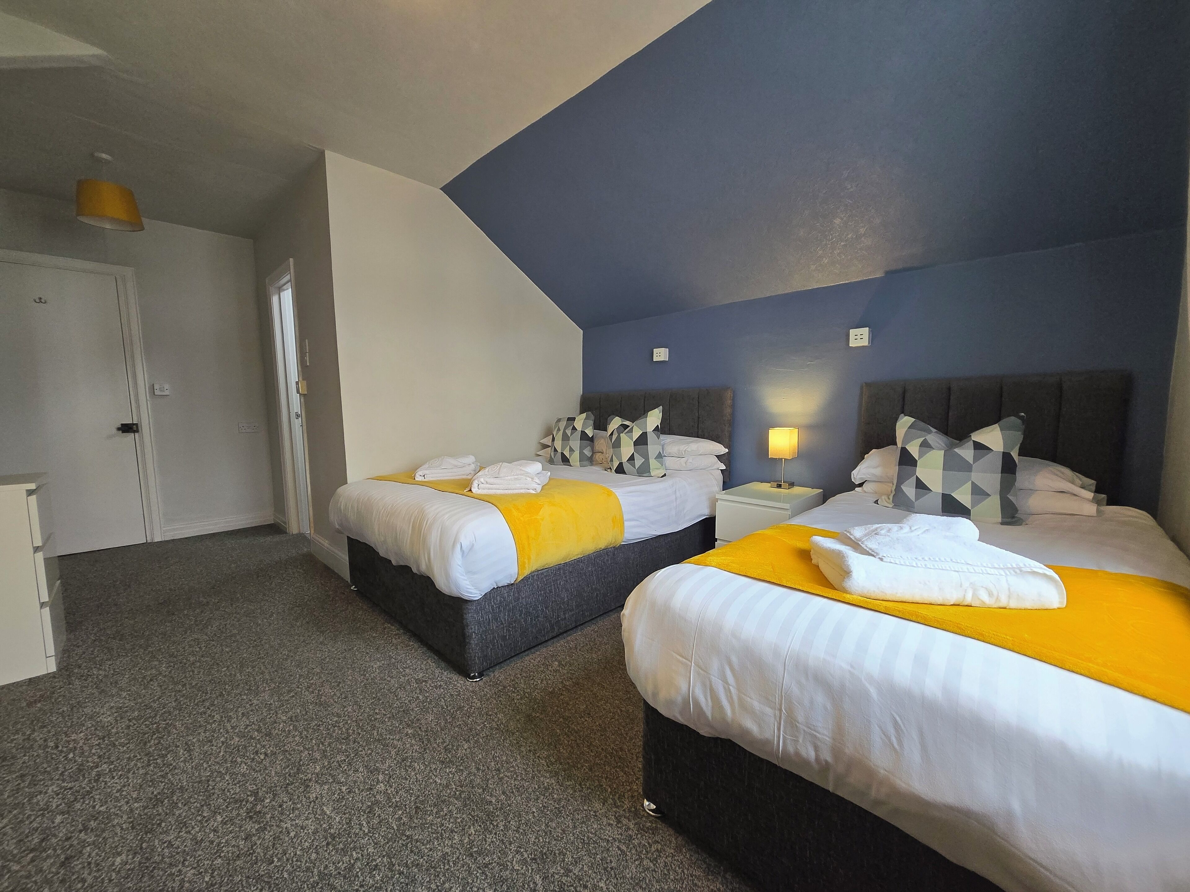 Une chambre d’hôtel avec deux lits, chacun recouvert d’une couverture jaune et de draps blancs, un tapis gris et une armoire blanche.