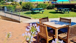An outdoor patio with wooden tables and chairs, picnic tables, and a view of a park with a clear blue sky. - Beach Hotel Torbay (Paignton)
