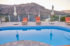 A swimming pool with lounge chairs and umbrellas, surrounded by a fence and a mountainous backdrop. - Best Family Friendly Villa Near Lake Kournas (Apokoronas)