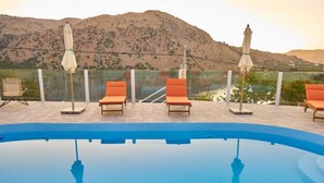 A swimming pool with lounge chairs and umbrellas, surrounded by a fence and a mountainous landscape. - Best Family Friendly Villa Near Lake Kournas (Apokoronas)