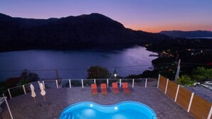 A swimming pool with lounge chairs and umbrellas on a terrace overlooking a lake at dusk. - Best Family Friendly Villa Near Lake Kournas (Apokoronas)