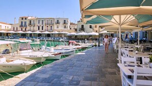 A marina with boats docked, a paved walkway, and outdoor seating areas with umbrellas. - Gorgeous Lake Kournas Villa Brand New Private Pool (Apokoronas)