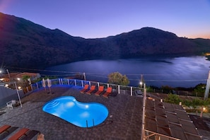 Une piscine entourée de chaises longues, elle-même bordée d’une terrasse dotée de panneaux solaires, offrant une vue sur un lac et des montagnes.