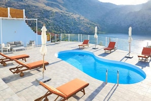 Un espace piscine doté de chaises longues et de parasols, offrant une vue sur un lac avec des montagnes en arrière-plan.