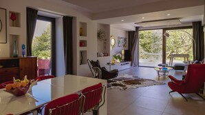 A modern living room with a dining area, large windows, and a view of greenery outside. - Alcantara Home view (Gravà - Castiglione di Sicilia)