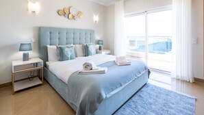 A modern bedroom with a large bed, a nightstand, a wall-mounted lamp, and a view of the outside through a large window. - Casa Malhotra at Boa Vista Golf Resort, Bayview Village (Lagos)