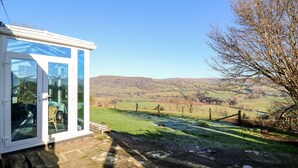 A conservatory with a glass door, a stone patio, and a view of rolling hills. - Ty Twmp / Tump Cottage (Brecon)