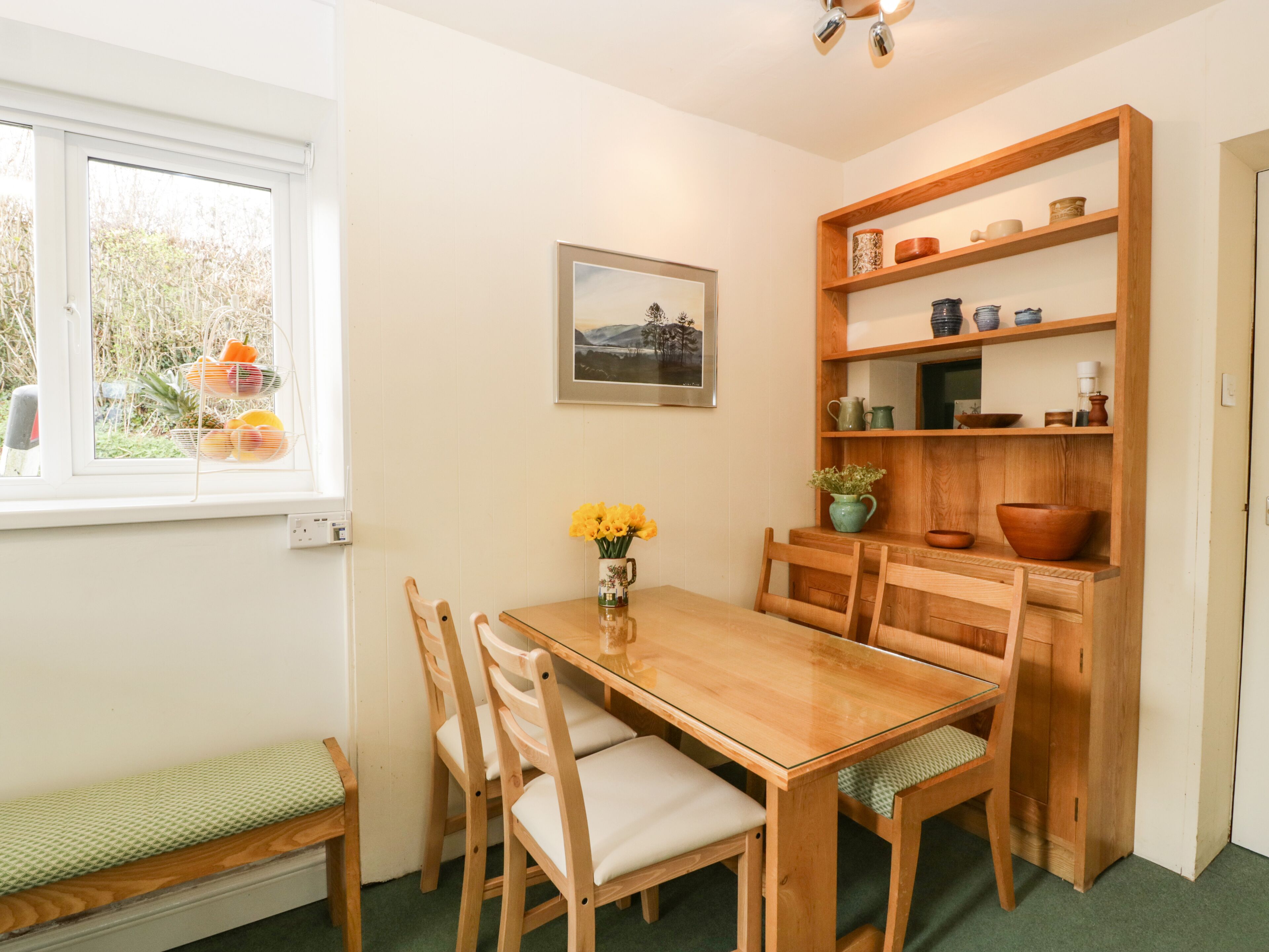 Un comedor con una mesa de madera, sillas, un banco, una estantería con utensilios de cocina, un cuadro enmarcado y una ventana con cestas de fruta.