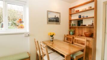 Un comedor con una mesa de madera, sillas, un banco, una estantería con utensilios de cocina, un cuadro enmarcado y una ventana con cestas de fruta.