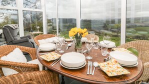 A conservatory dining area with a table set for four, surrounded by wicker chairs, and large windows offering a view of the countryside. - Ty Twmp / Tump Cottage (Brecon)