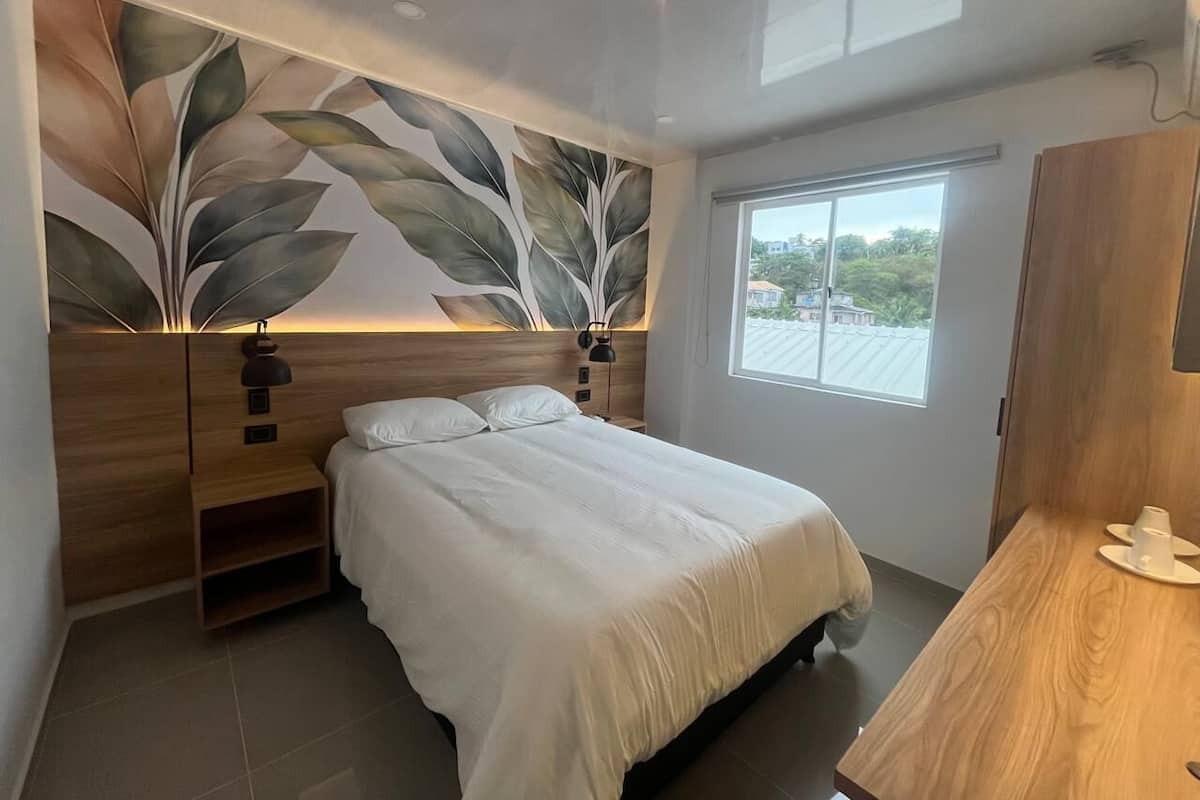 A bedroom with a bed, a nightstand, a wall-mounted lamp, and a window.