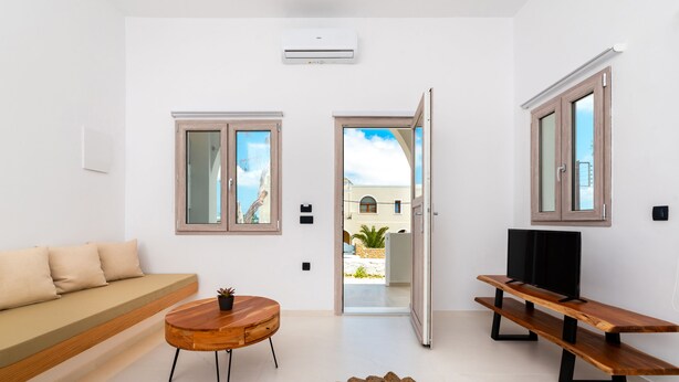 A modern living room with a sofa, a wooden coffee table, a TV on a wooden stand, and a view through an open door to an outdoor area.
