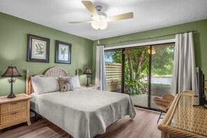 Ein Schlafzimmer mit einem Bett, zwei Nachttischen, einem Deckenventilator, einem Fenster mit Vorhängen und Blick ins Grüne.