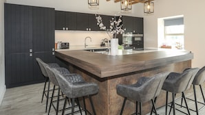 A modern kitchen with dark cabinets, a wooden island with bar stools, pendant lights, and a visible oven.