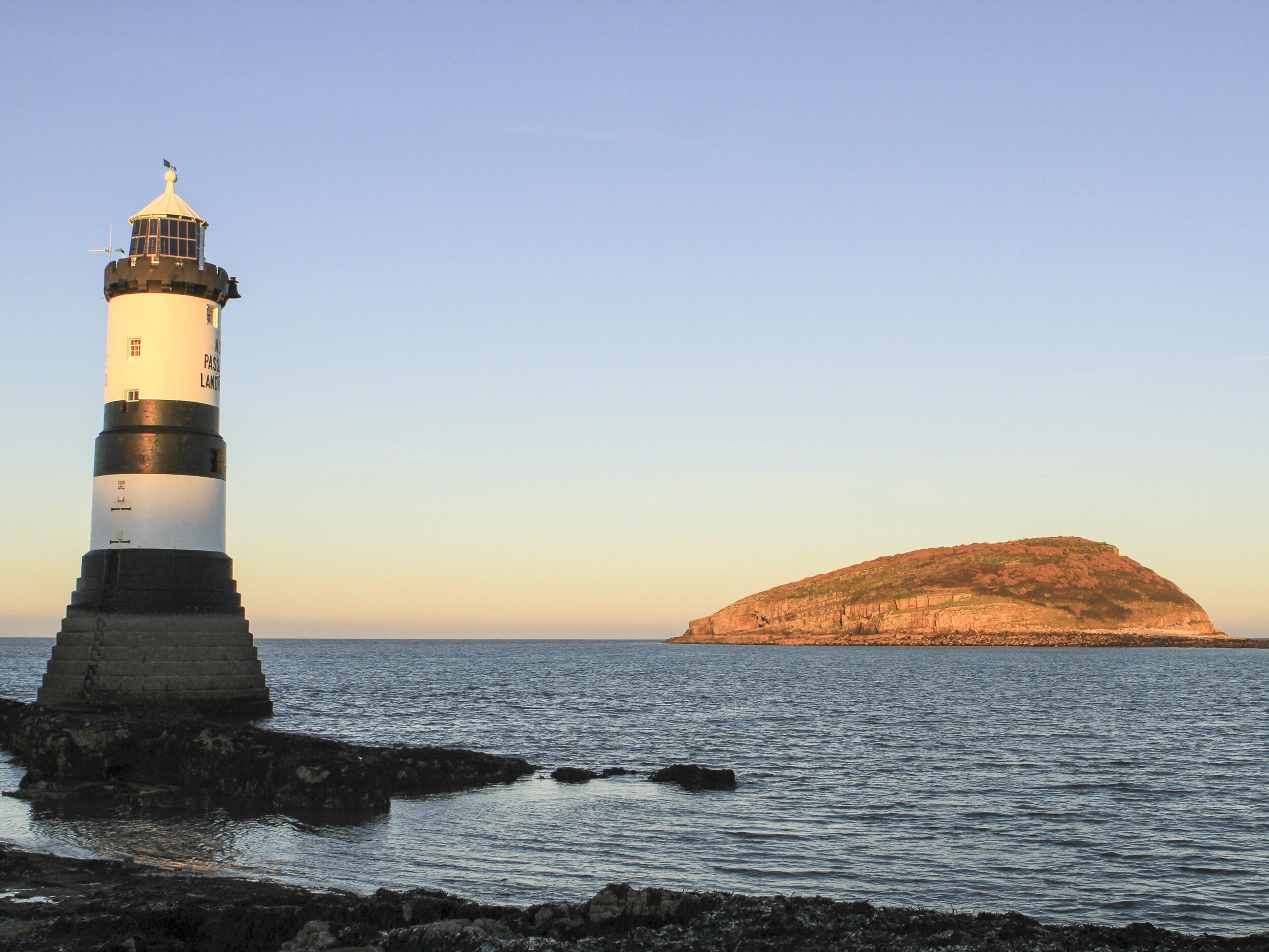 Um farol sobre um afloramento rochoso, com águas calmas e uma ilha ao longe.