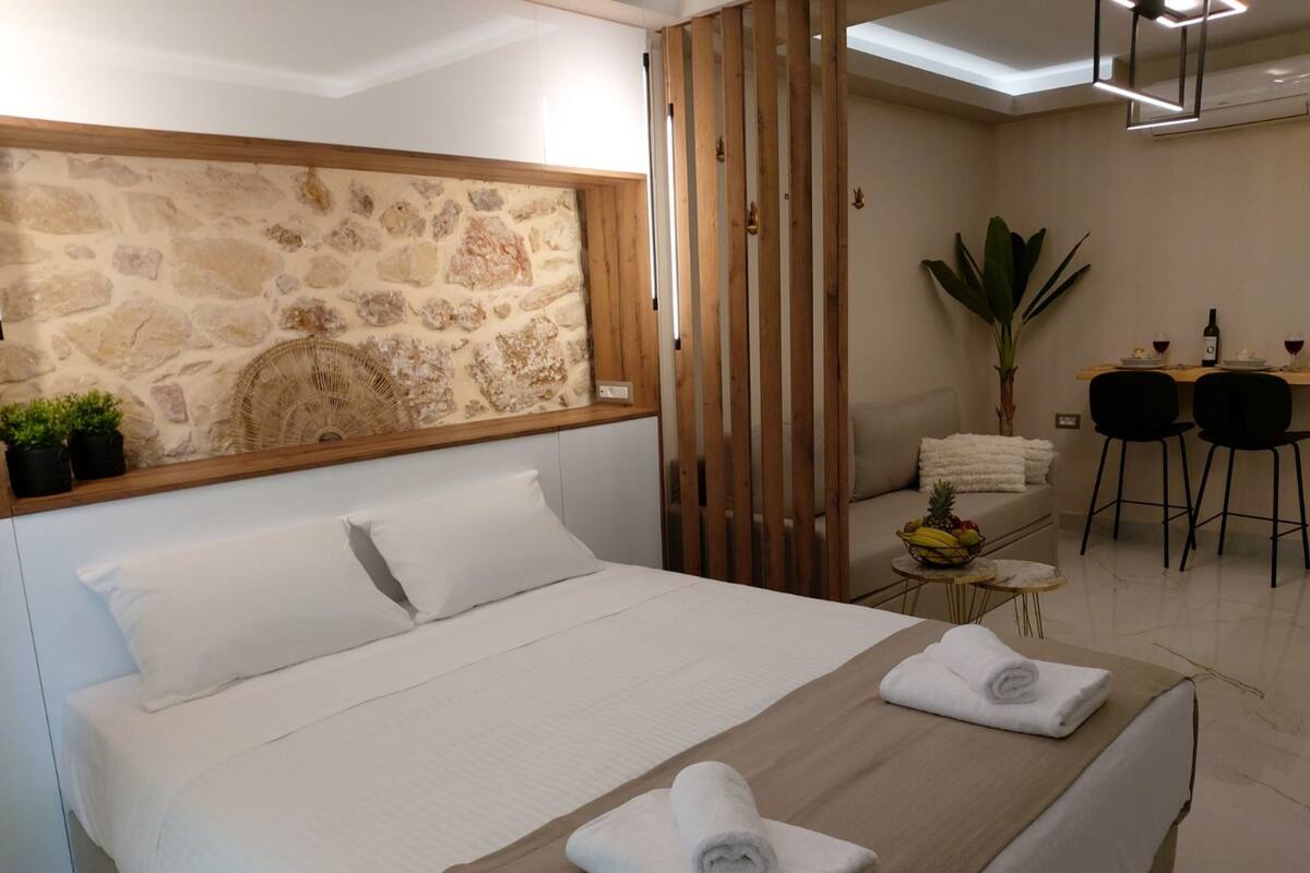 Habitación de hotel con pared de acento de piedra, cama con ropa de cama blanca, separador de madera, pequeña mesa con frutero y zona de comedor con sillas y planta.