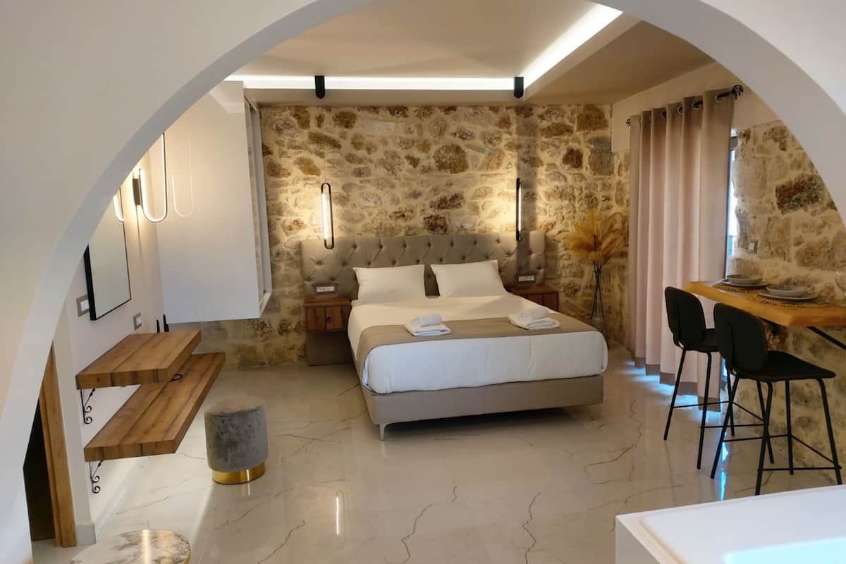 Un dormitorio moderno con pared de piedra, cama con cabecera capitonada, escritorio de madera y zona de estar con sillones.
