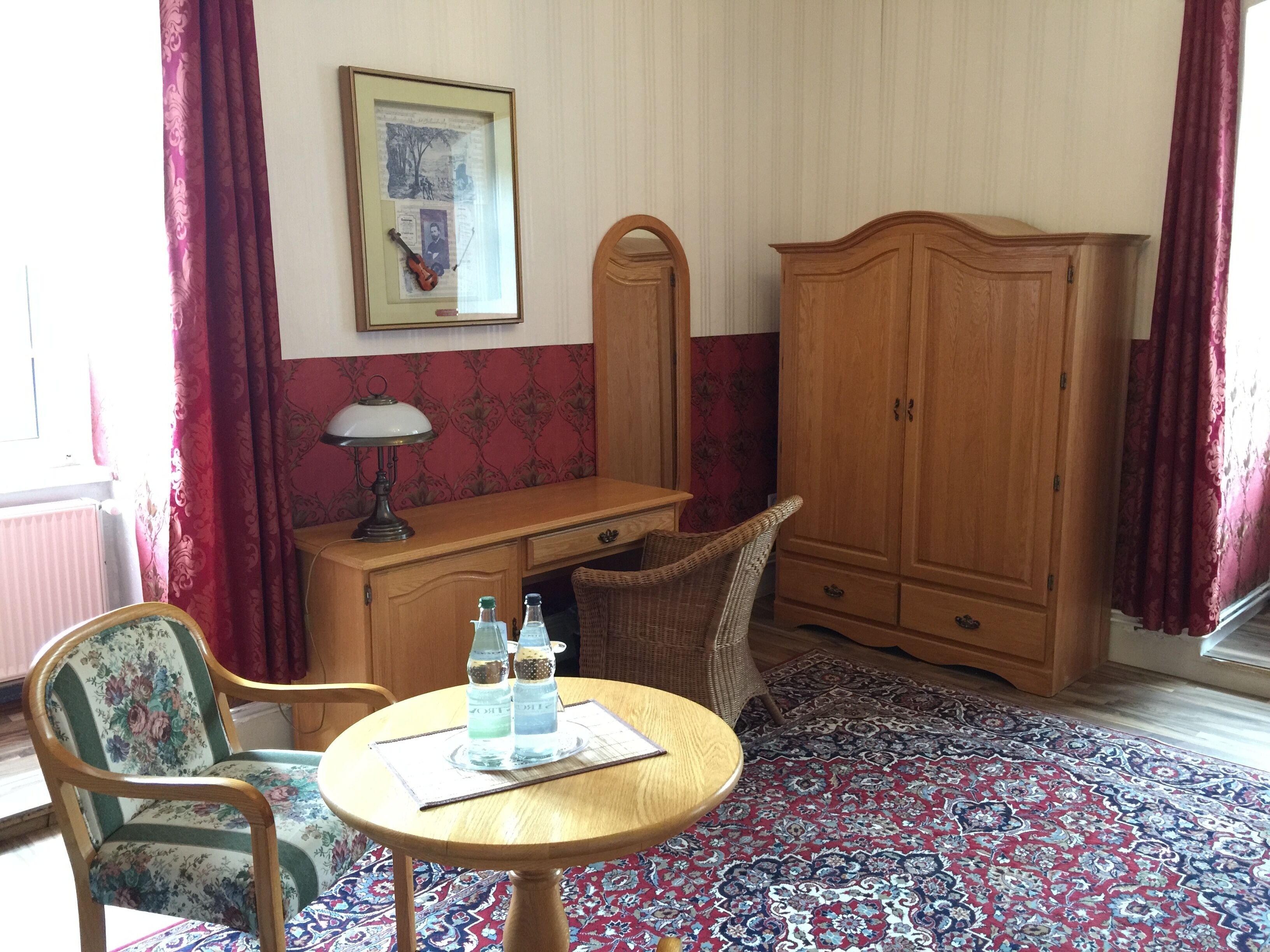 A room with a wooden desk, a chair, a table with bottles, a mirror, and a patterned rug.