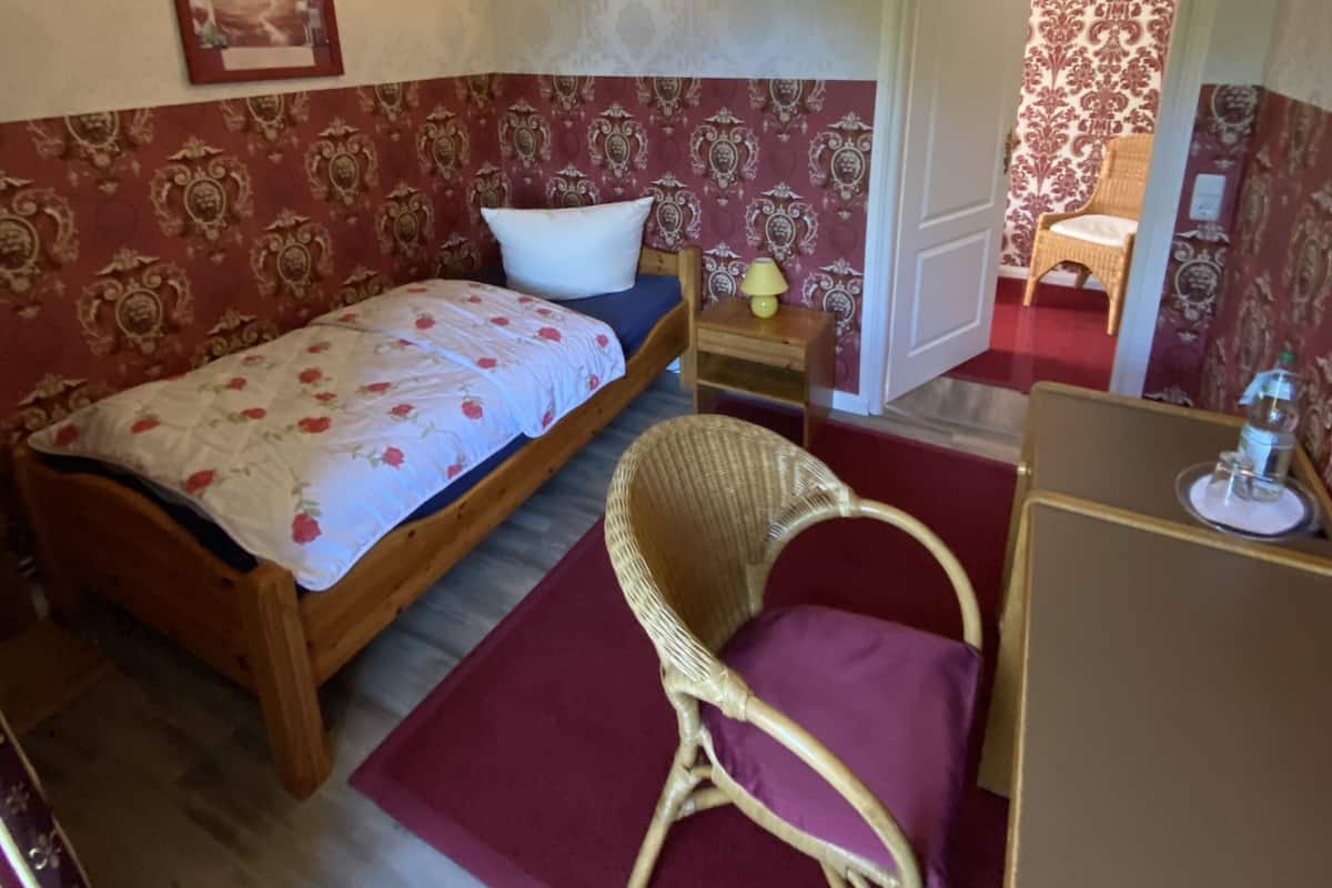A bedroom with a wooden bed, a wicker chair, a small table with a lamp, and a door leading to another room.