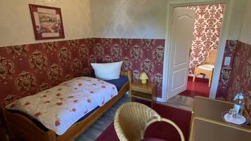 A bedroom with a wooden bed, a wicker chair, a small table with a lamp, and a door leading to another room.
