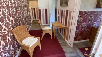 Un pasillo estrecho con papel tapiz estampado, alfombra roja, muebles de mimbre y una pequeña mesa con objetos decorativos.