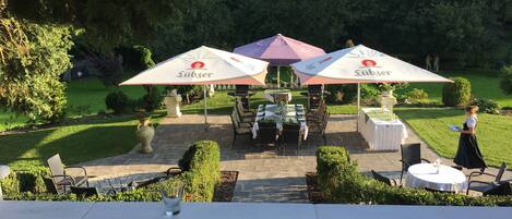 Een terras met tafels en stoelen onder grote parasols, omgeven door groen en een verzorgd gazon.