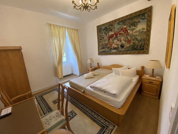 A bedroom with a double bed, two bedside tables with lamps, a wooden chair, a desk, a radiator, and a large painting on the wall. - Jagdschloss Lalendorf (Lalendorf)