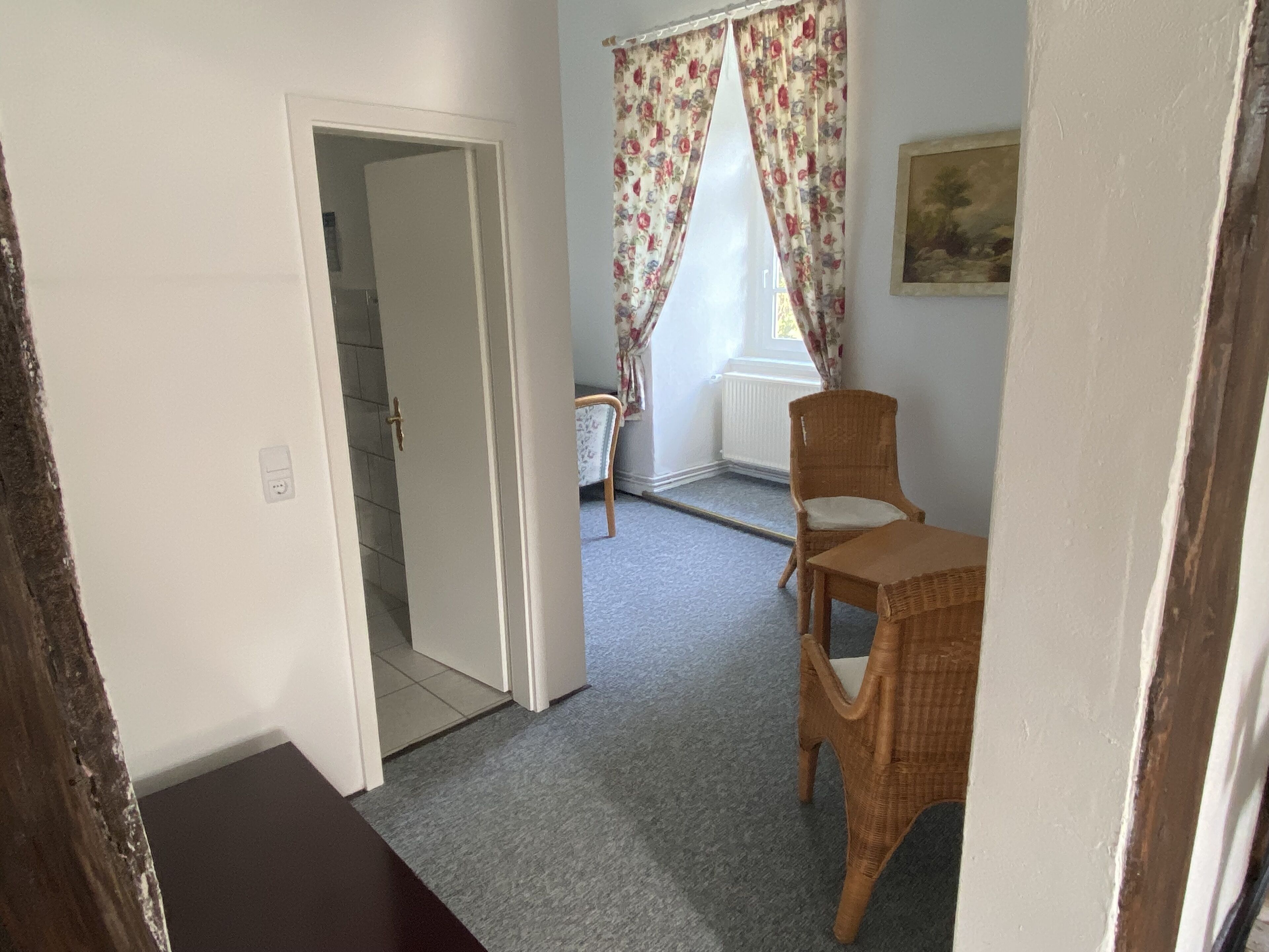 A room with a door leading to another space, a wicker chair, a small table, and a window with floral curtains.
