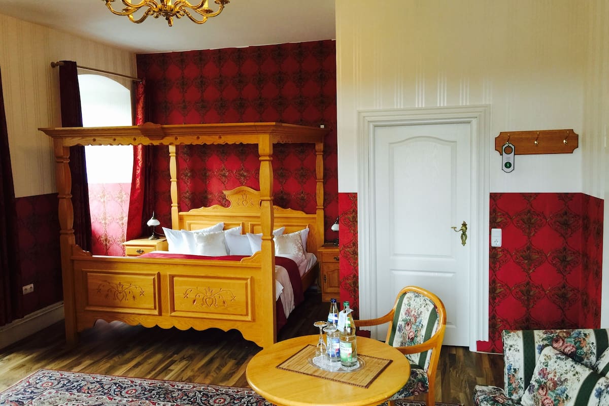 A four-poster bed with a wooden headboard, a round wooden table with a placemat, and a floral-patterned armchair.