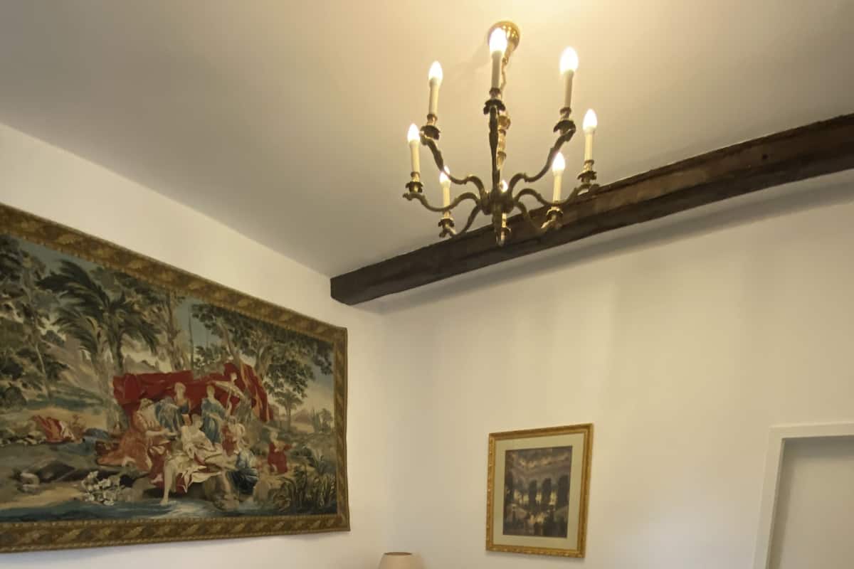 A double bed with white linens, a nightstand with a lamp, a large tapestry, a framed picture, and a door.