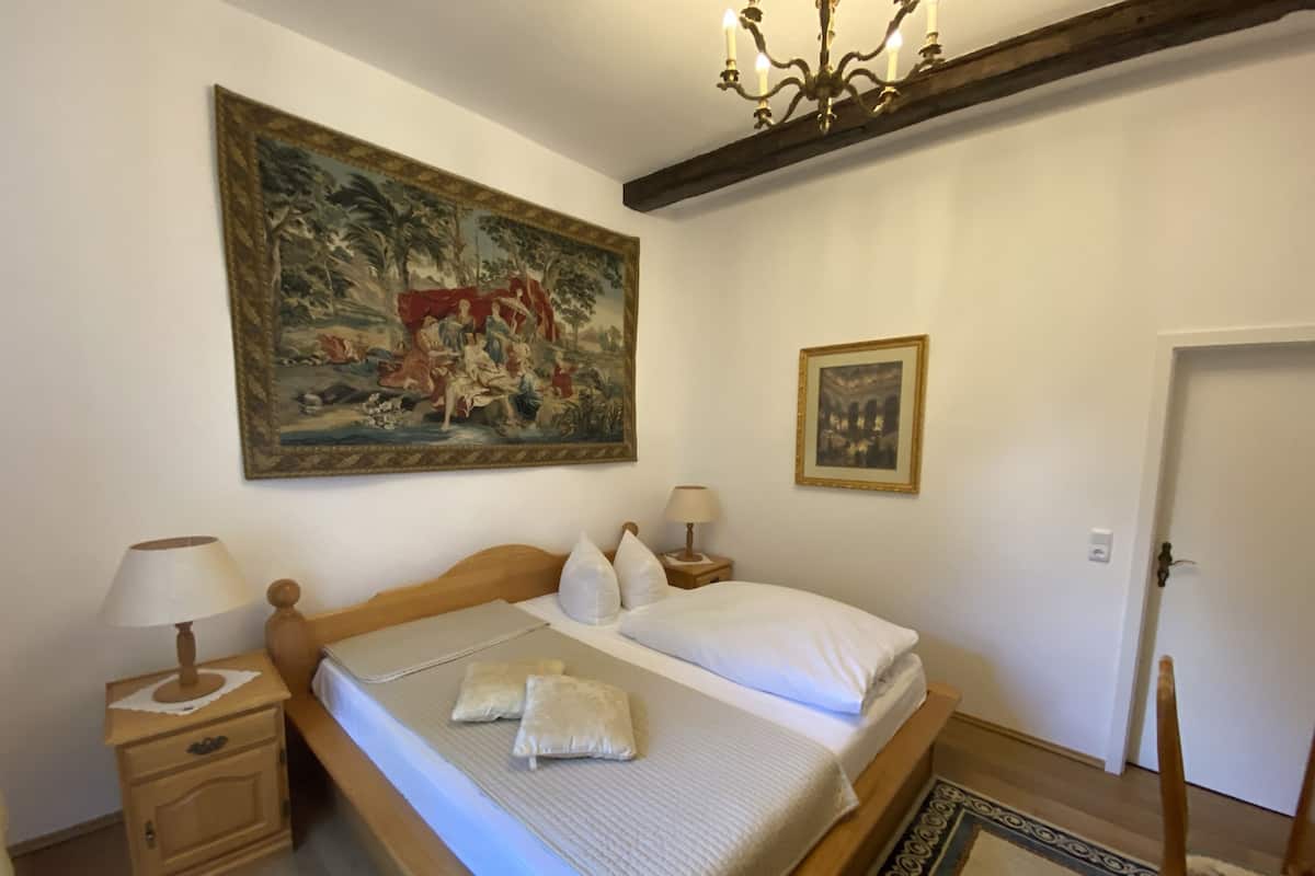 A bedroom with a bed, a nightstand, a lamp, a painting, and a chandelier.