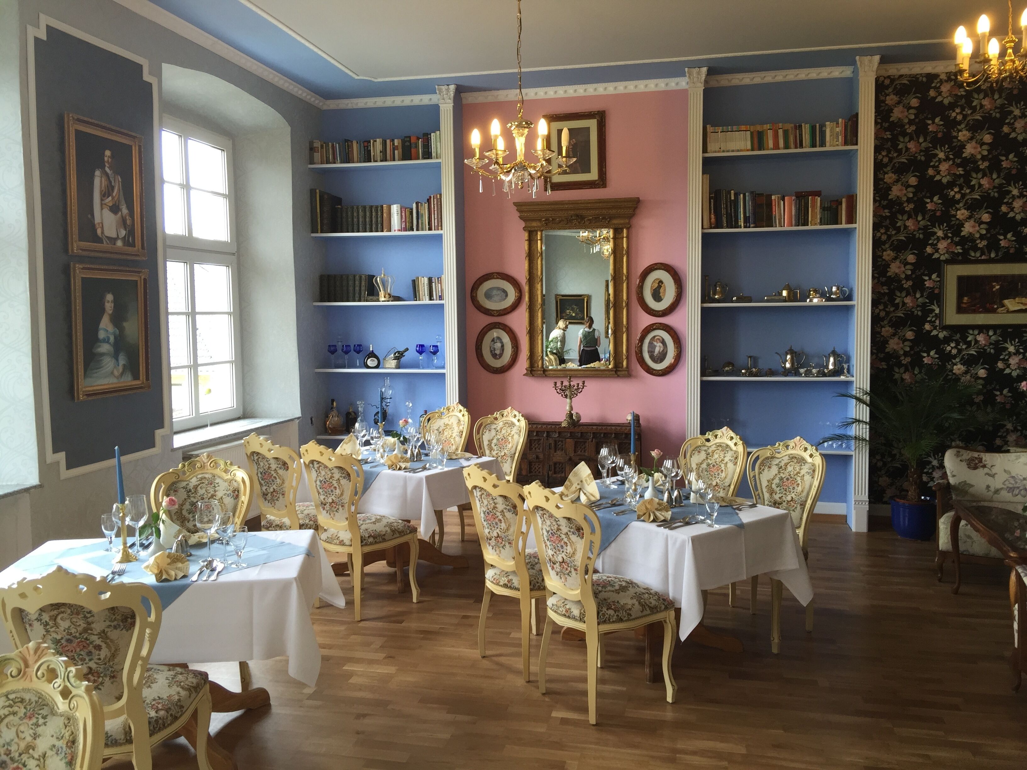 A dining room with ornate chairs, a chandelier, and a bookshelf filled with books.