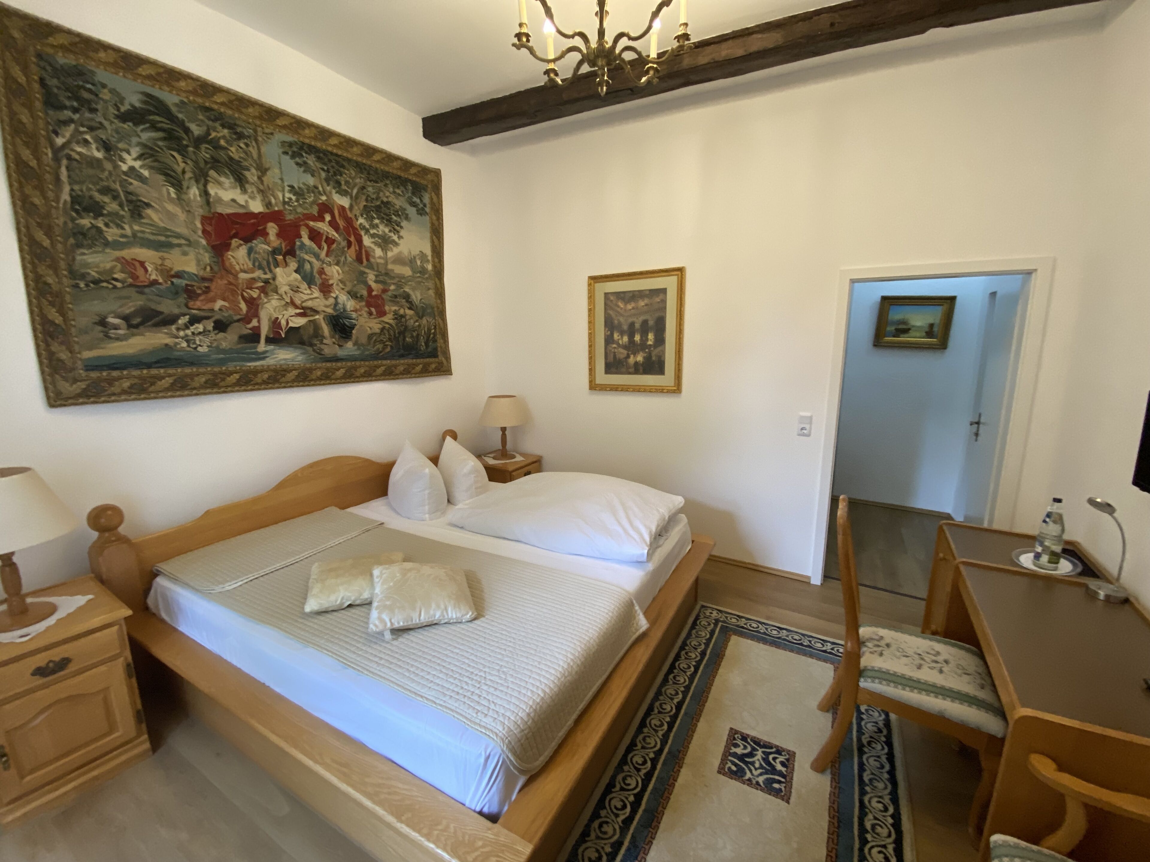 Un dormitorio con cama de madera, ropa de cama blanca, un cuadro grande, una mesita de noche con lámpara y una puerta.